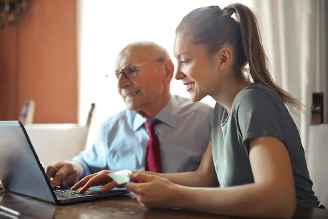 A girl holding a credit card in front of a laptop with an elderly person